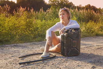 sad girl sits on a country road with a fuel tank and a hose waiting for help in the rays of the sunset. Toned. Portrait