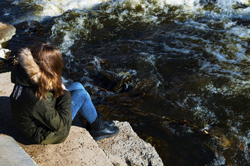 Young girl sitting on the river bank, looking at the raging water.