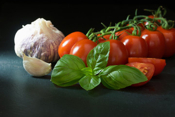 Low key shot of shrubs tomatoes with a leaf of basil, garlic, clove of garlic and sliced tomato lying on the black stone table. 