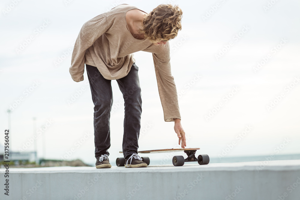 Wall mural young curly long haired man riding on longboard on empty road