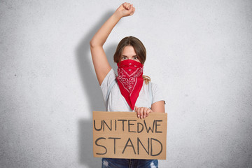 Isolated shot of strong female shows women power, raises hand, shows fists, holds plate with inscription, defenses all females, wears red bandana on face, isolated over white studio background