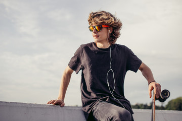 Young hipster man standing near concrete wall with longboard using his phone and headphones