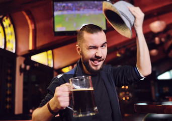 a man in a Bavarian hat drinking beer in a pub and watching football on a monitor during Oktoberfest