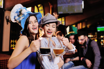 friends - boys and girls in Bavarian hats drinking beer on the background of the bar during the celebration of Oktoberfest or St. Patrick's Day