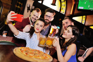group or company of young people - friends drink beer, eat pizza, talk and laugh and shoot selfie on the smartphone's camera on the background of the bar