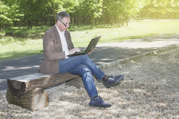 Fototapeta premium A grown man in a brown jackets sits with an open laptop on a wooden bench in a summer park