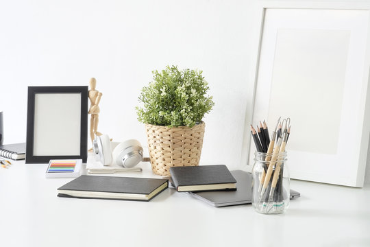 Artist workplace table with jar of pencil, sketch book, photo frame and plant decoration on white table.