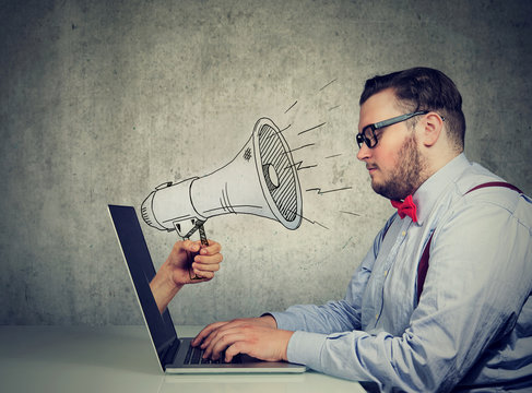 man sitting at table working on computer screaming with a megaphone poking out from a laptop screen