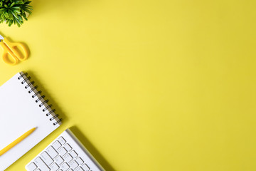 Above Office desk with computer keyboard, notebook paper, pen and plant decoration on yellow background top view mock up.