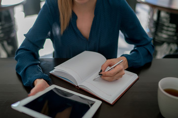 Cropped view of woman writes information in notebook while calculating month budget and all outgoing costs using tablet. Selective focus on student's hand with pen noting items of learning material