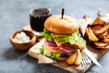 Tasty grilled beef burger with bacon, pickled onion and french fries served on a rustic wooden board on dark slate background