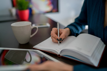Selective focus on notepad with female hand writing down notepad pages, woman blogger recording creative ideas for article sitting with modern digital tablet, notebook planner and cup of coffee