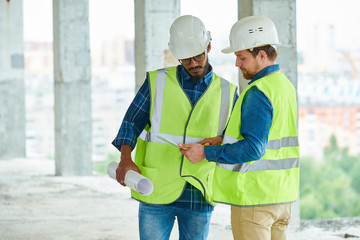 Two young men in hardhats and waistcoats looking at scheme on tablet while working on construction site together