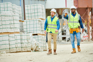 Two young men in hardhats and waistcoats pointing at distance while walking near stacks of building...