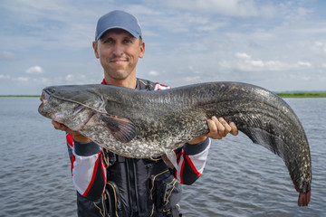 Happy fisherman with big catfish trophy at the boat with fishing tackles