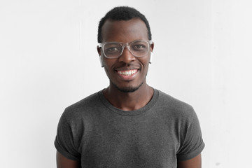 Horizontal shot of young dark-skinned man with positive smile on face, isolated on gray background, wearing plastic glasses, being in high spirit because of personal success, dressed in casual T-shirt