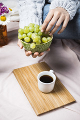 Woman's hands in soft sweater holding wooden bowl with green grapes with cup of black coffee on wooden tray, selective focus