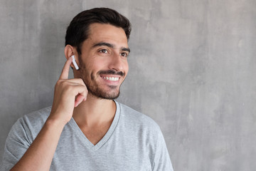 Young man wearing wireless earbuds and t shirt, listening to his favorite musical album online, touching one earphone to control application - obrazy, fototapety, plakaty