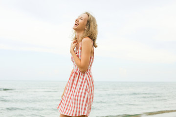 Fototapeta na wymiar Young beautiful woman with blondie hair is laughing with toothy smile, wearing short red checked dress on seashore