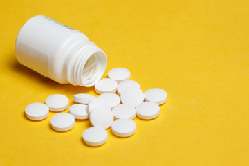 White pills spilling out of a white plastic pill bottle on light yellow background