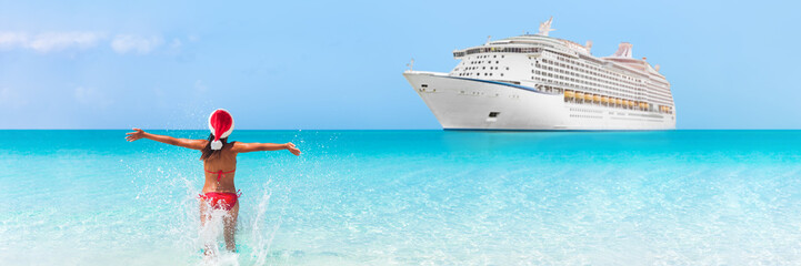 Christmas cruise travel vacation holidays in Caribbean beach. Happy bikini woman wearing santa claus hat on new year holiday running in happiness in blue turquoise water with ship in background. - obrazy, fototapety, plakaty