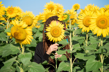 girl with dreadlocks is shy and hiding behind a sunflower in summer on the field. Hippie plays hide...