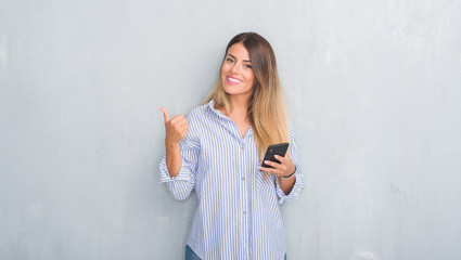 Young adult woman over grey grunge wall looking at smartphone texting a message happy with big smile doing ok sign, thumb up with fingers, excellent sign