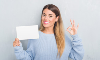 Naklejka premium Young adult woman over grey grunge wall holding blank card doing ok sign with fingers, excellent symbol