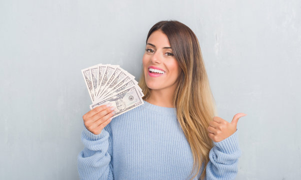 Young adult woman over grey grunge wall holding dollars pointing and showing with thumb up to the side with happy face smiling