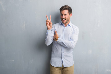 Handsome young business man over grey grunge wall wearing elegant shirt smiling with happy face winking at the camera doing victory sign. Number two.