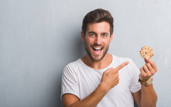 Handsome Young Man Over Grey Grunge Wall Eating Chocolate Chip Cooky Very Happy Pointing With Hand And Finger