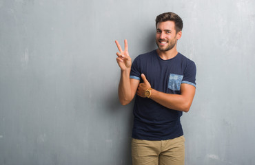Handsome young man over grey grunge wall smiling with happy face winking at the camera doing victory sign. Number two.