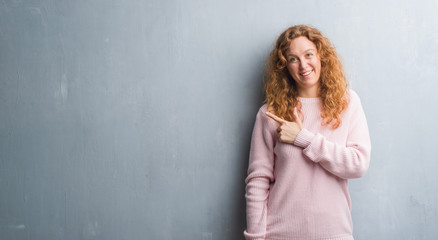 Fototapeta premium Young redhead woman over grey grunge wall wearing pink sweater cheerful with a smile of face pointing with hand and finger up to the side with happy and natural expression on face.