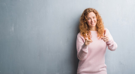 Young redhead woman over grey grunge wall wearing pink sweater pointing fingers to camera with happy and funny face. Good energy and vibes.