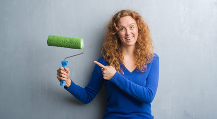 Young redhead woman over grey grunge wall holding painting roller very happy pointing with hand and finger