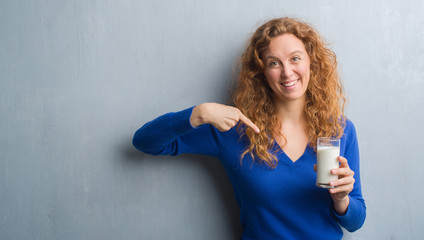 Young redhead woman drinking glass of milk with surprise face pointing finger to himself