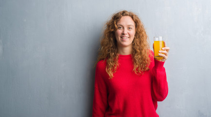 Young redhead woman over grey grunge wall drinking a glass of orange juice with a happy face standing and smiling with a confident smile showing teeth