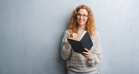 Young redhead woman over grey grunge wall reading a book happy with big smile doing ok sign, thumb up with fingers, excellent sign