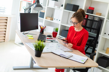 A young girl is sitting at the desk in the office, holding a bank card and phone in her hand.