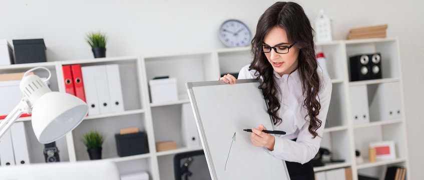 A beautiful young girl stands near an office desk and draws a magnetic marker on the magnetic board.