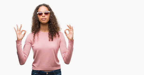 Beautiful young hispanic woman wearing sunglasses relax and smiling with eyes closed doing meditation gesture with fingers. Yoga concept.