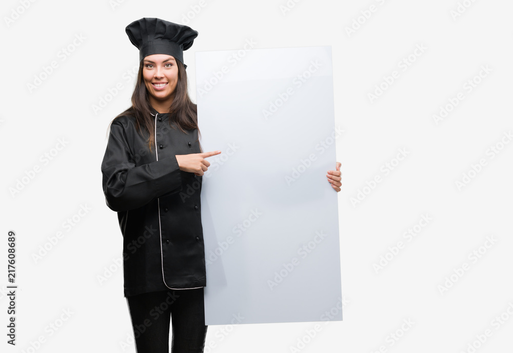 Wall mural young hispanic cook woman wearing chef uniform holding banner very happy pointing with hand and fing