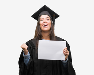 Young hispanic woman wearing graduated uniform holding diploma paper screaming proud and celebrating victory and success very excited, cheering emotion