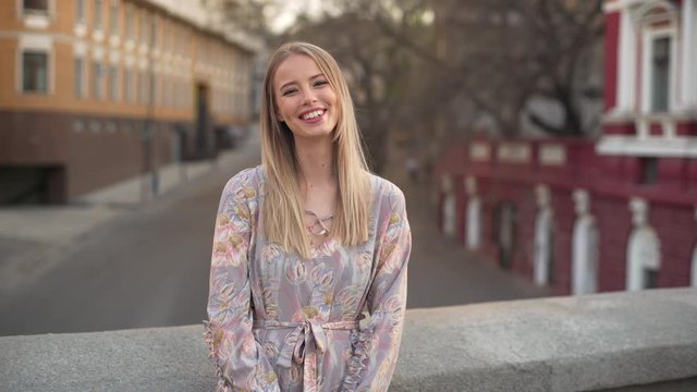 Portrait of excited young woman 20s in beautiful dress laughing, while walking outdoor with cars and buildings