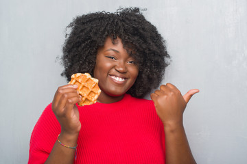 Young african american woman over grey grunge wall eating belgium waffle pointing and showing with thumb up to the side with happy face smiling