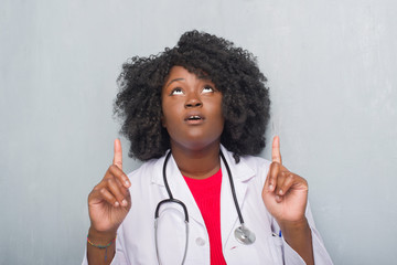 Young african american doctor woman over grey grunge wall amazed and surprised looking up and pointing with fingers and raised arms.