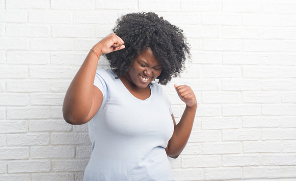 Young african american plus size woman over white brick wall very happy and excited doing winner gesture with arms raised, smiling and screaming for success. Celebration concept.