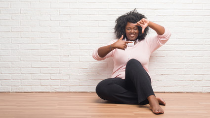 Young african american woman sitting on the floor wearing headphones smiling making frame with hands and fingers with happy face. Creativity and photography concept.