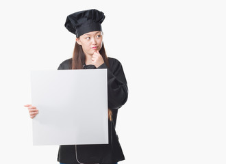 Young Chinese woman over isolated background wearing chef uniform holding banner serious face thinking about question, very confused idea