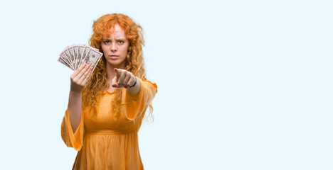 Young redhead woman holding dollars pointing with finger to the camera and to you, hand sign, positive and confident gesture from the front